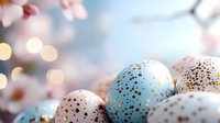 Speckled Easter eggs in pastel blue and brown tones with gold dots against blurred spring cherry blossom background with soft bokeh lights.