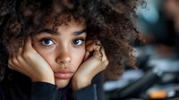 Young African American woman with natural curly hair and striking blue eyes resting face on hands, looking thoughtfully at camera against blurred background.