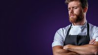 Young male chef with ginger beard wearing black apron and white uniform stands confidently with crossed arms against dramatic purple background, professional portrait.