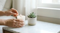 Female hands lighting white candle on marble windowsill next to small succulent plant in ceramic pot creating peaceful home atmosphere for relaxation and mindfulness.