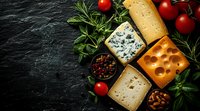 Various cheese types with blue cheese, emmental, and aged cheddar arranged on dark stone background with fresh herbs, cherry tomatoes and mixed peppercorns.