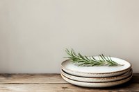 Stack of handmade ceramic plates with rosemary sprig on wooden surface against neutral beige wall background for kitchen and dining decor.