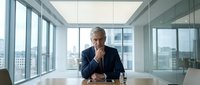 Mature male executive in dark suit sitting at conference table in modern high-rise office with city skyline views, contemplative expression, professional business environment.