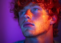 Young male with freckles and curly ginger hair in dramatic purple and orange neon lighting looking up, showing defined facial features in creative portrait photography.