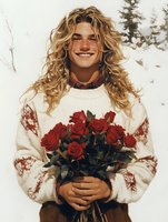 Young Caucasian man with long blonde wavy hair wearing white knit sweater holds bouquet of red roses, expressing genuine joy and warmth against winter background.