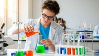 Young female scientist in lab coat and glasses conducting chemical experiment with colorful solutions in modern laboratory setting.