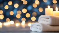 Glowing candles on folded white spa towels against blurred warm bokeh lights background creating relaxing atmosphere for wellness and meditation.