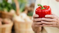 Red bell pepper held in hands of mature adult with blurred fresh produce in background, representing organic farming, healthy eating, and farm-to-table food selection.