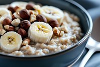 Creamy oatmeal bowl topped with fresh banana slices and hazelnuts in dark ceramic bowl, healthy breakfast closeup with selective focus on morning meal.