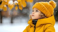 Adorable toddler girl in yellow winter coat and knit hat looking up outdoors in snowy park with autumn leaves. Child winter fashion and seasonal activities.