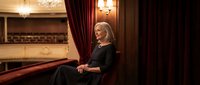 Elegant senior woman in black dress sitting in luxurious opera house theater box with red velvet curtains and ornate interior for cultural events.