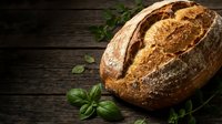 Freshly baked artisan sourdough bread loaf with golden crust on rustic wooden table with fresh basil herbs for bakery and cooking concepts.