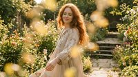 Young redhead woman in flowing beige dress walking through blooming garden path with warm sunlight and bokeh effects creating dreamy romantic atmosphere.
