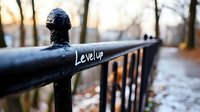 Black metal railing with level up text graffiti in urban park setting during sunset, blurred background with walking people and autumn trees creates motivational scene.