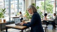 Senior woman with gray hair and glasses working on laptop at wooden desk in modern office space with large windows, potted plants, and colleagues in background during daytime.