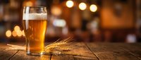 Golden beer glass with wheat stalks on rustic wooden table in warm pub atmosphere with bokeh lights creating cozy drinking establishment ambiance.