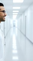 Male healthcare professional wearing glasses and white coat standing in modern hospital corridor with fluorescent lighting.