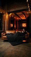 Woman relaxing on tan leather couch in industrial loft with neon sign, warm string lights, and exposed brick walls. Urban interior design and lifestyle.