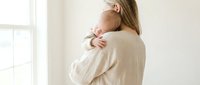 Young Caucasian mother gently holds sleeping newborn baby on her shoulder near bright window, soft natural light, tender motherhood moment.