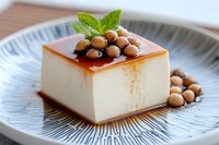Square slice of silky smooth pudding topped with caramel sauce, roasted macadamia nuts and fresh mint leaf garnish served on blue ceramic plate.