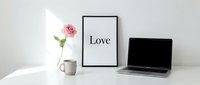 Minimalist home office desk with laptop, pink rose, coffee mug, and love quote poster on white background for Valentine's Day.