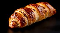 Fresh baked golden croissant with glossy surface and flaky layers on black background, dramatic lighting highlights buttery texture and crispy exterior.