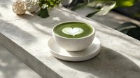 Green matcha latte with heart-shaped foam art in white ceramic cup on concrete surface, natural sunlight casting soft shadows through leaves.