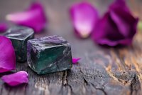 Raw fluorite crystal cubes in deep green color with purple rose petals scattered on rustic wooden surface, soft focus macro photography with bokeh effect.