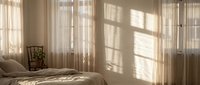 Sunlit bedroom with warm natural light streaming through sheer curtains creating geometric shadows on neutral bedding and wooden floors in minimalist interior.
