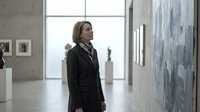 Young woman with long brown hair, wearing a black blazer and dark clothing, standing in a contemporary art gallery or museum space viewing abstract artwork on white walls.
