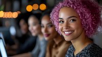 Young woman with vibrant pink curly hair smiling warmly while working in modern office environment with diverse coworkers and soft bokeh lights in background.