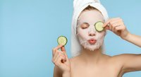 Young Caucasian woman with white towel on head applying cucumber slices on eyes during facial skincare routine with cream mask on blue background.