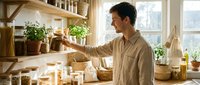 Middle-aged woman with dark hair and olive skin tone, wearing a beige linen shirt, organizing eco-friendly products and plants on wooden shelves in a bright, minimalist kitchen space with natural light.