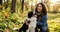 Young woman with curly brown hair embracing black and white border collie dog in sunny autumn park with golden light filtering through trees.