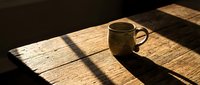 Ceramic mug with hot beverage sitting on wooden table in natural sunlight, casting long shadows. Suitable for coffee shop, lifestyle, and morning routine content.