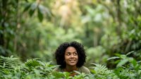 Young woman with curly dark hair and medium skin tone smiling among lush green foliage in natural outdoor setting, suitable for wellness, nature, lifestyle and environmental campaigns.