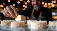 Caucasian male chef in black apron garnishing desserts with powdered sugar against blurred bokeh lights background, close up of hands working.