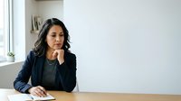 Thoughtful woman with dark wavy hair, South Asian ethnicity, wearing navy blazer and gray top, sitting at desk with hand to face in contemplative pose, suitable for business and professional concepts.