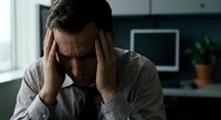 Stressed middle-aged Caucasian businessman holding head in hands experiencing headache or mental fatigue in modern office workspace for wellness concepts.