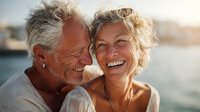 Senior Caucasian couple laughing together at sunset by the sea, showcasing genuine happiness and romantic connection in their golden years.