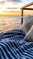 Cozy striped blanket and beige pillow on outdoor lounge chair at sunset, with golden ocean horizon and dramatic sky creating peaceful vacation atmosphere.