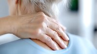 Person massaging tense neck muscles to relieve pain and stiffness, close-up view of hands on shoulder against blurred background in medical setting.