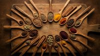 Wooden spoons arranged in circle filled with colorful spices, herbs, seeds and seasonings on rustic wood background for cooking and culinary projects.
