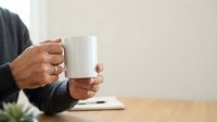 Adult male with medium skin tone holding blank white mug at wooden desk with notebook and plant, wearing dark sweater. Suitable for branding, mockups, and beverage marketing campaigns.