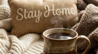 Cozy home atmosphere with steaming ceramic mug of hot tea or coffee, knitted blanket, and Stay Home pillow in warm beige tones.