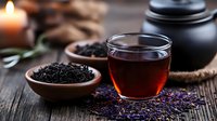 Aromatic black tea in glass cup with loose tea leaves in wooden bowl, burning candle and teapot on rustic table with lavender flowers for relaxation.