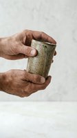 Elderly hands holding handmade ceramic mug with speckled glaze against neutral background for artisan pottery and craftsmanship concepts.