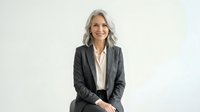 Mature woman with long gray hair, Caucasian ethnicity, wearing charcoal blazer and white blouse, smiling confidently against white background for corporate and business use.