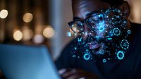 African American male professional wearing glasses illuminated by blue digital interface projections while working on laptop in dark environment with bokeh lights.