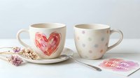 Ceramic mugs with heart and polka dot designs on white background, paired with saucers, spoon, dried flowers and pink gift tag for Valentine's Day, Mother's Day, or home decor styling.